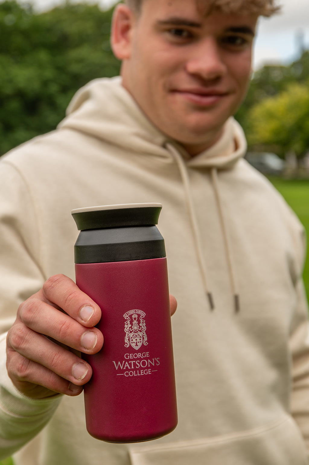 A person in a beige hoodie holds up a dark red insulated water bottle with a black lid. The bottle features the George Watson's College crest and name printed in white.