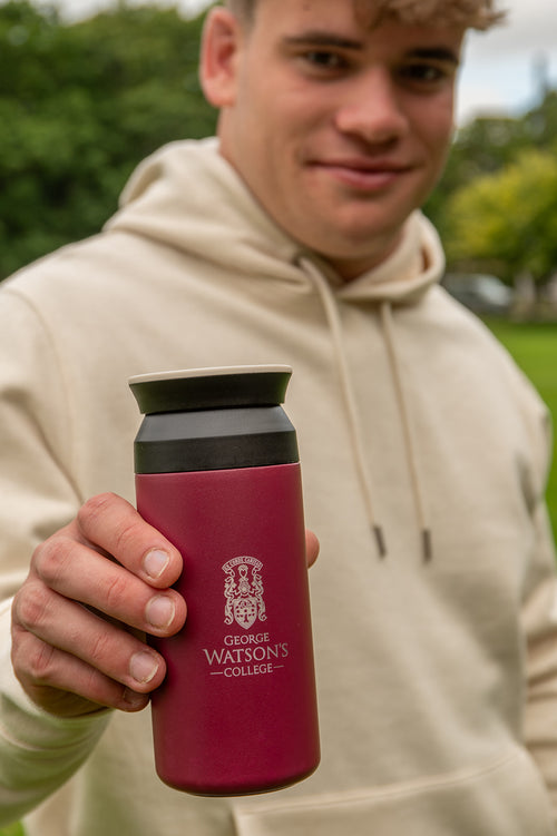A person in a beige hoodie holds up a dark red insulated water bottle with a black lid. The bottle features the George Watson's College crest and name printed in white.
